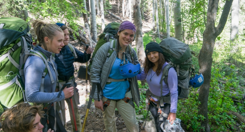 A group of students wearing backpacks and smile while standing in a shady area with green trees. 